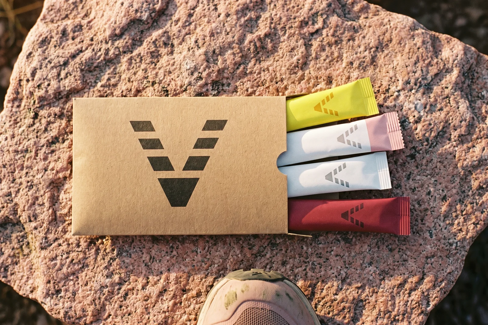A flat kraft sleeve printed with the VitaminHill mark on pink granite, four marked stick packs sliding out of it, yellow, white with a pink end, white and red, and the toe of a trail shoe at the edge.