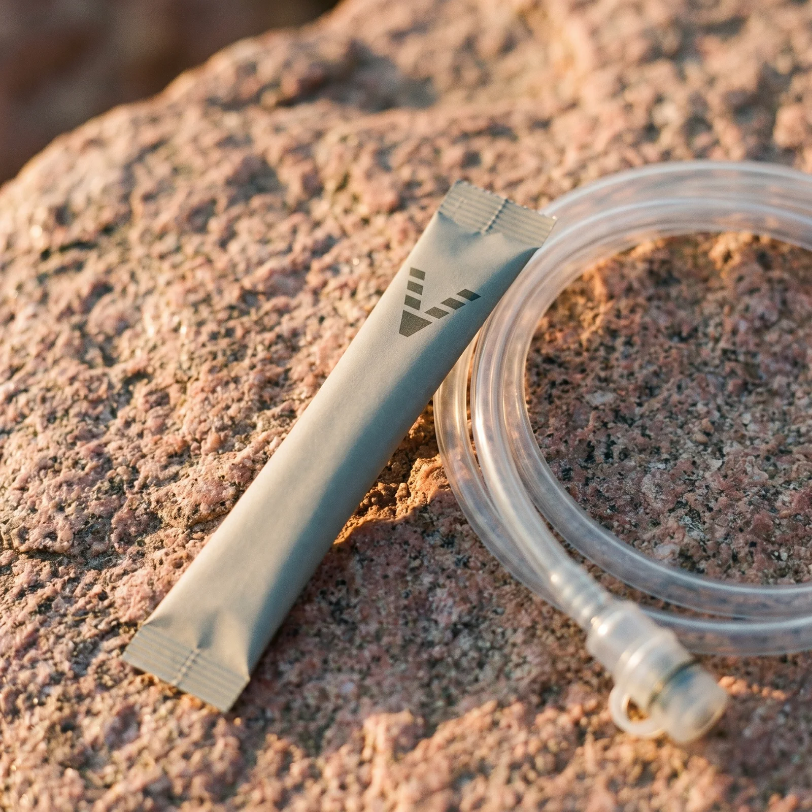 A slim stone-grey stick pack printed with the VitaminHill mark, on pink granite beside the coiled clear drinking tube of a hydration bladder.
