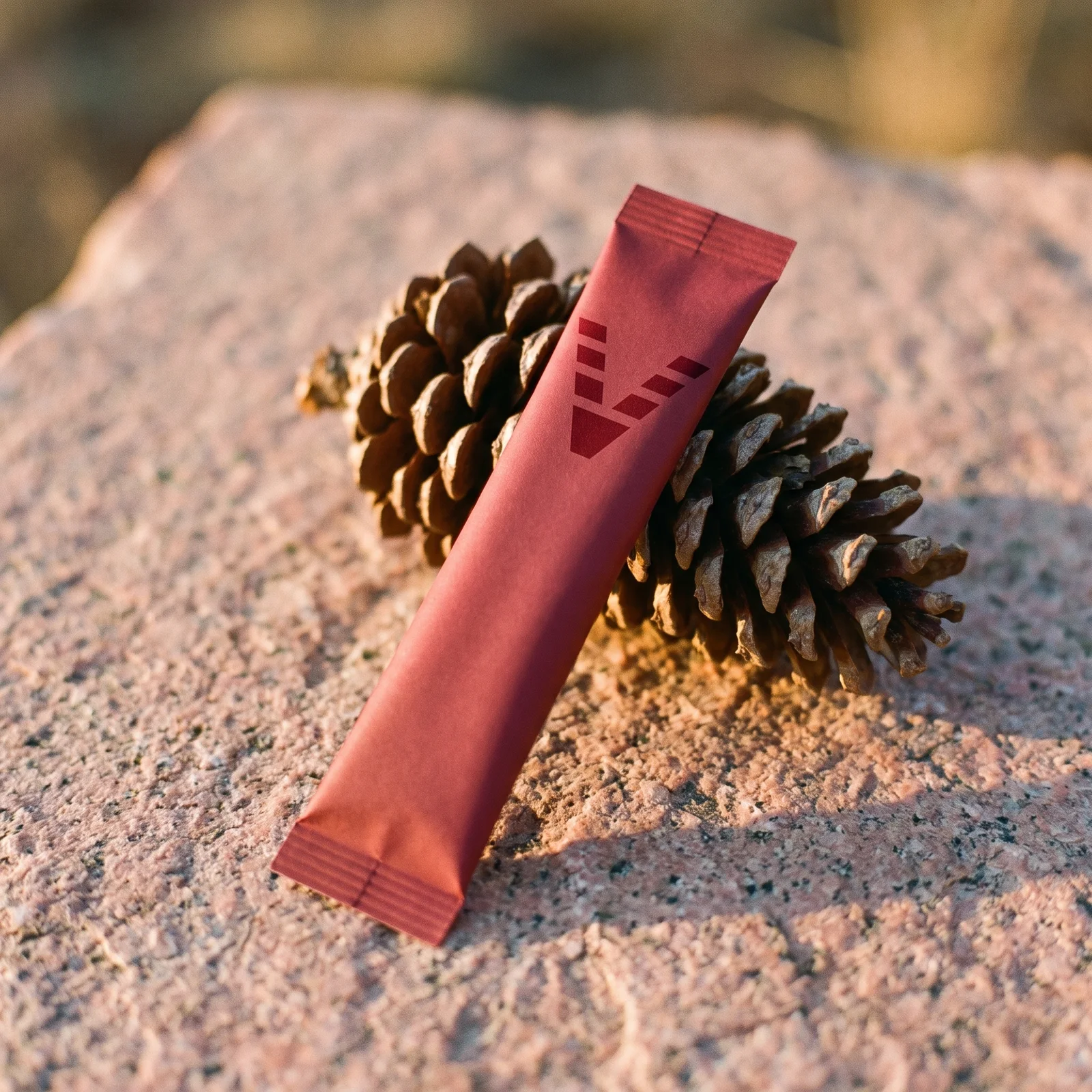 A slim tart-cherry red stick pack printed with the VitaminHill mark, leaning on a fallen ponderosa pine cone on pink granite.