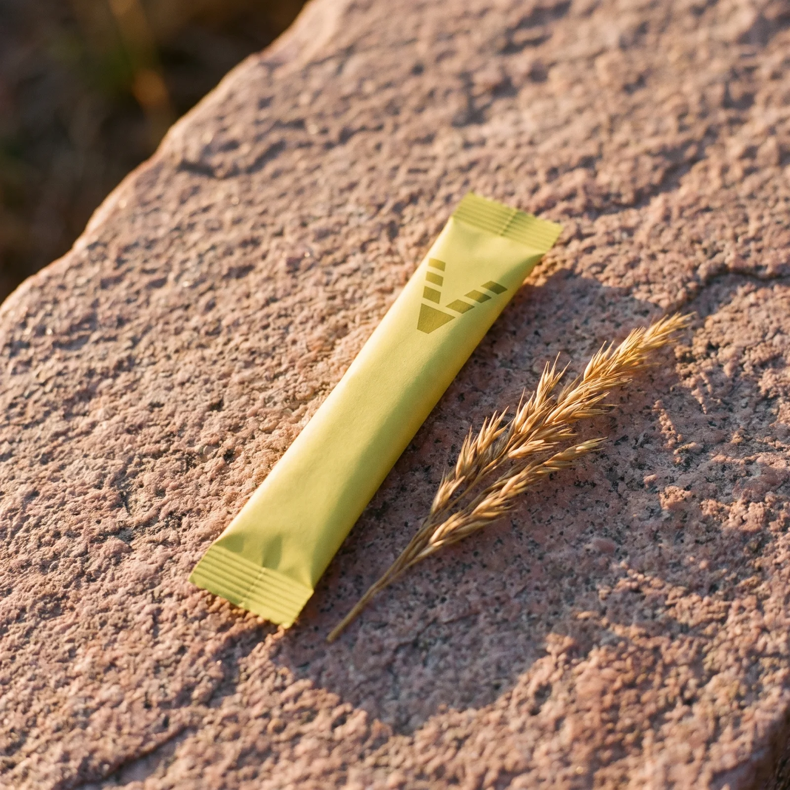 A slim citrus-yellow stick pack printed with the VitaminHill mark, lying across pink granite beside a sprig of dry grass.