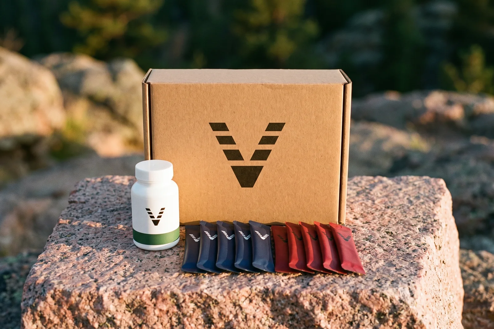 A kraft carton printed with the VitaminHill mark holding a white bottle with a green band, midnight blue sticks and red sticks, on pink granite.