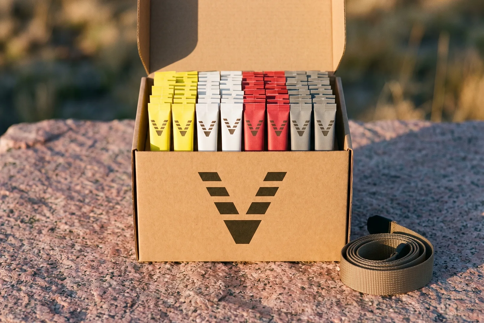 An open kraft box with the VitaminHill mark on its front, filled with rows of marked stick packs in four colors, on pink granite beside a coiled trekking-pole strap.