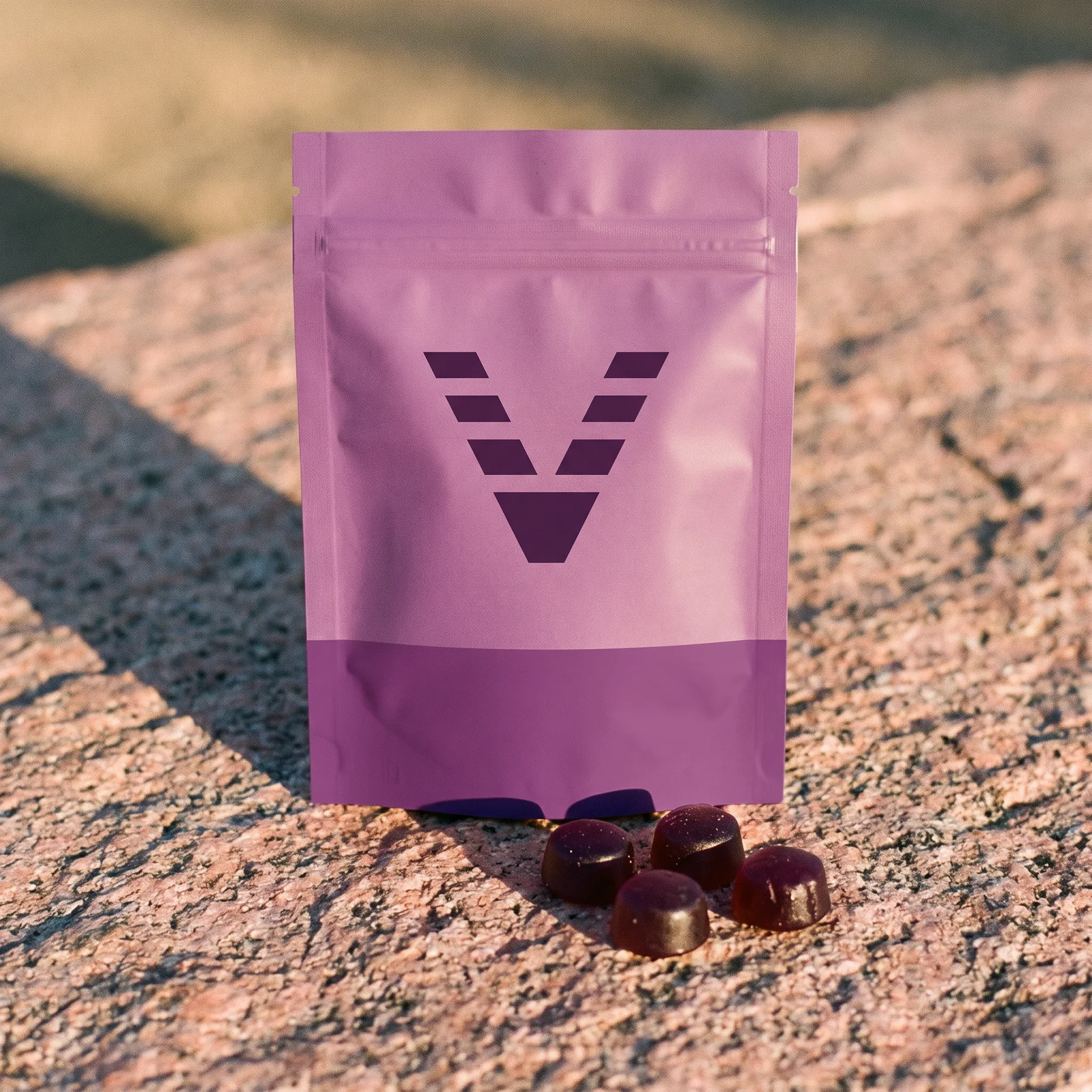 A flat matte chew pouch with a huckleberry purple band and the VitaminHill mark, on pink granite in late sun, a few purple chews beside it.
