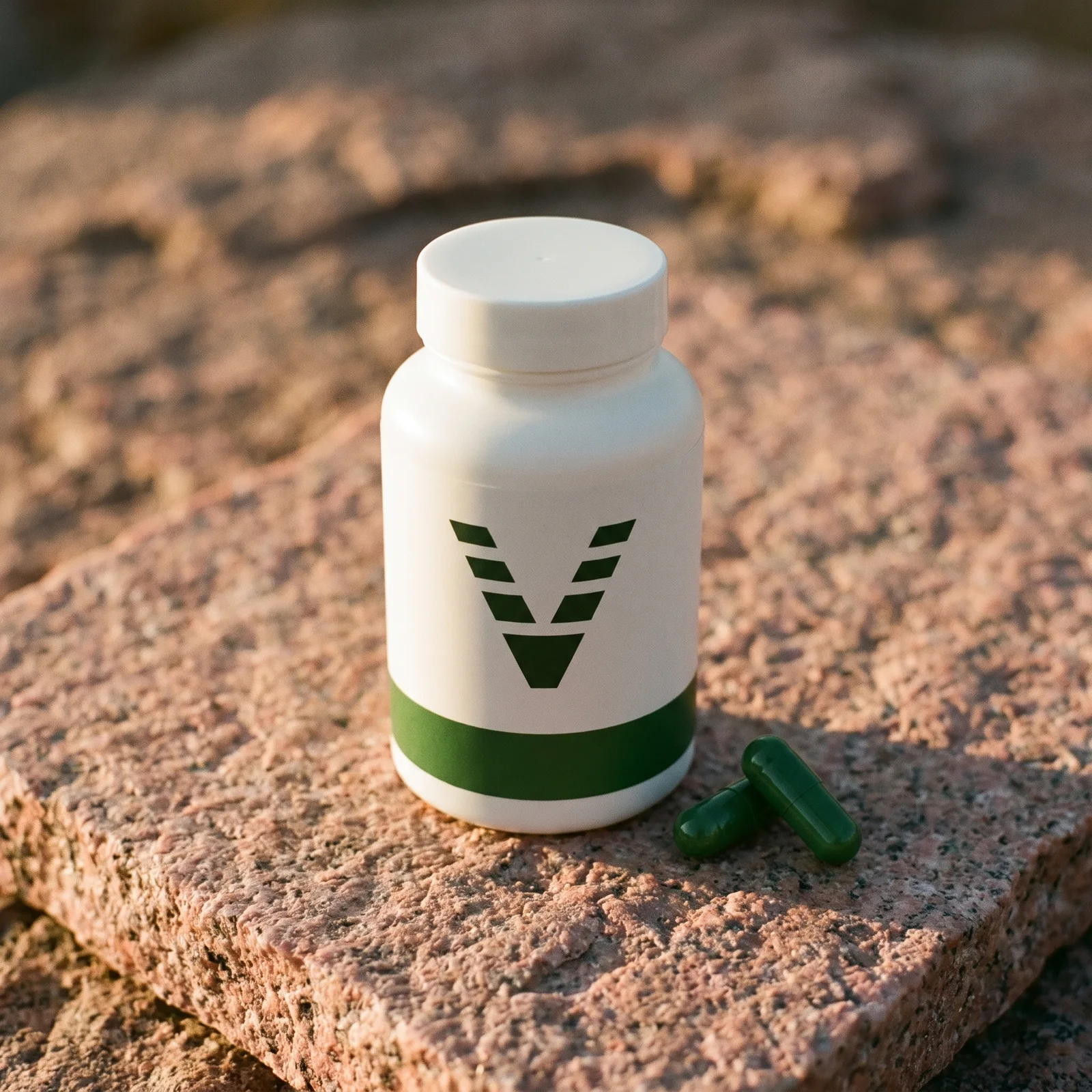 A matte white wide-mouth bottle with a forest green band and the VitaminHill mark, on pink granite, two green capsules beside it.