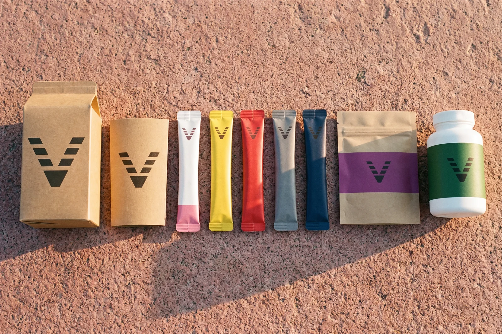 The whole VitaminHill line on pink granite in late sun: a kraft carton, stick packs in five colors, a purple chew pouch and a white bottle with a green band.